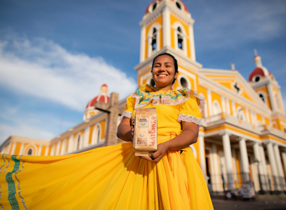 Woman in Indigenous dress holds a bag of Mayorga coffee beans in front of colonial building
