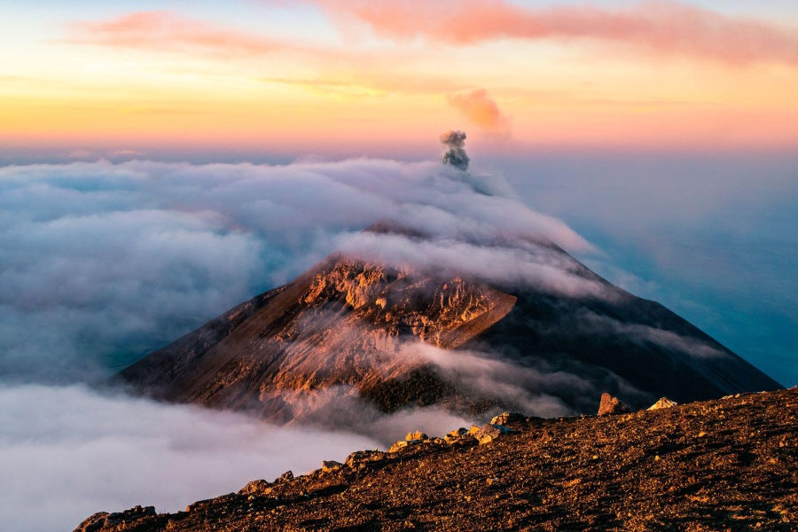 A volcano at sunrise, peeking through the clouds and emitting a puff of smoke