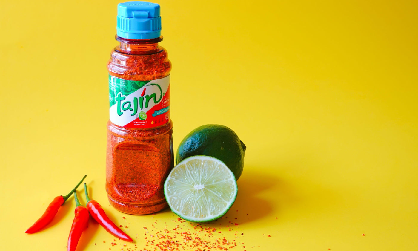 A shaker pot of tajín against a yellow backdrop with chiles to the left of it and limes to the right