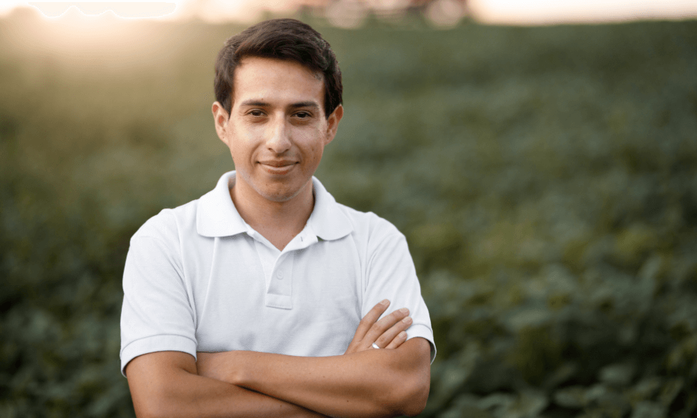 A Latino man stands in a field, arms folded and smiling