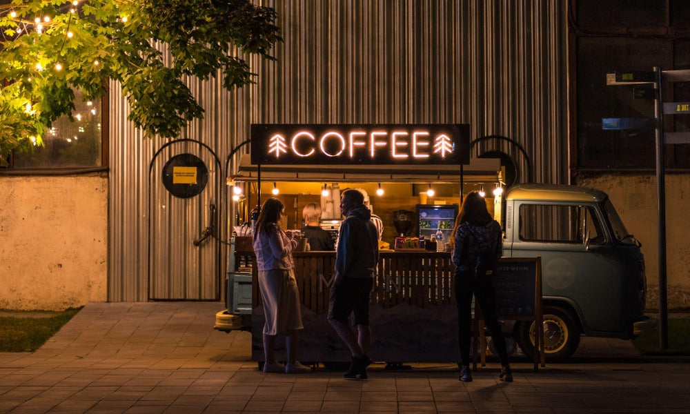 A coffee truck on the side of the road at nighttime with 3 people stood drinking coffee