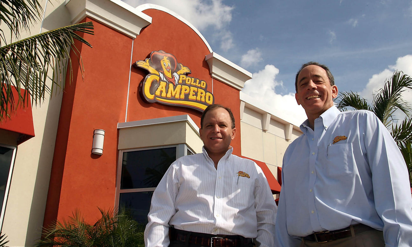 Two employees wearing Pollo Campero shirts stand outside a Pollo Campero restaurant