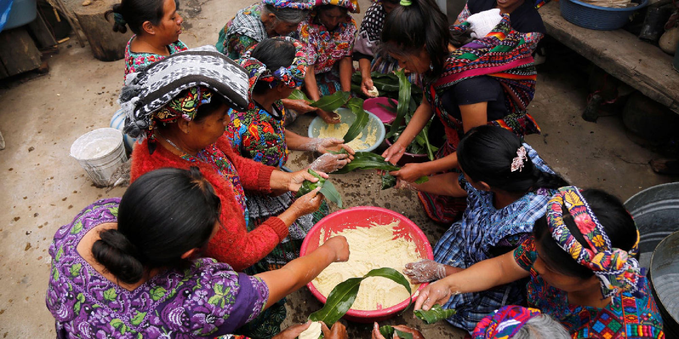 Women in indigenous dress make masa, crowded around a table together