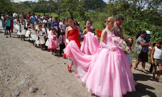 A girl walks down a country road with flocks of people and flower-girls behind her for her quinceañera